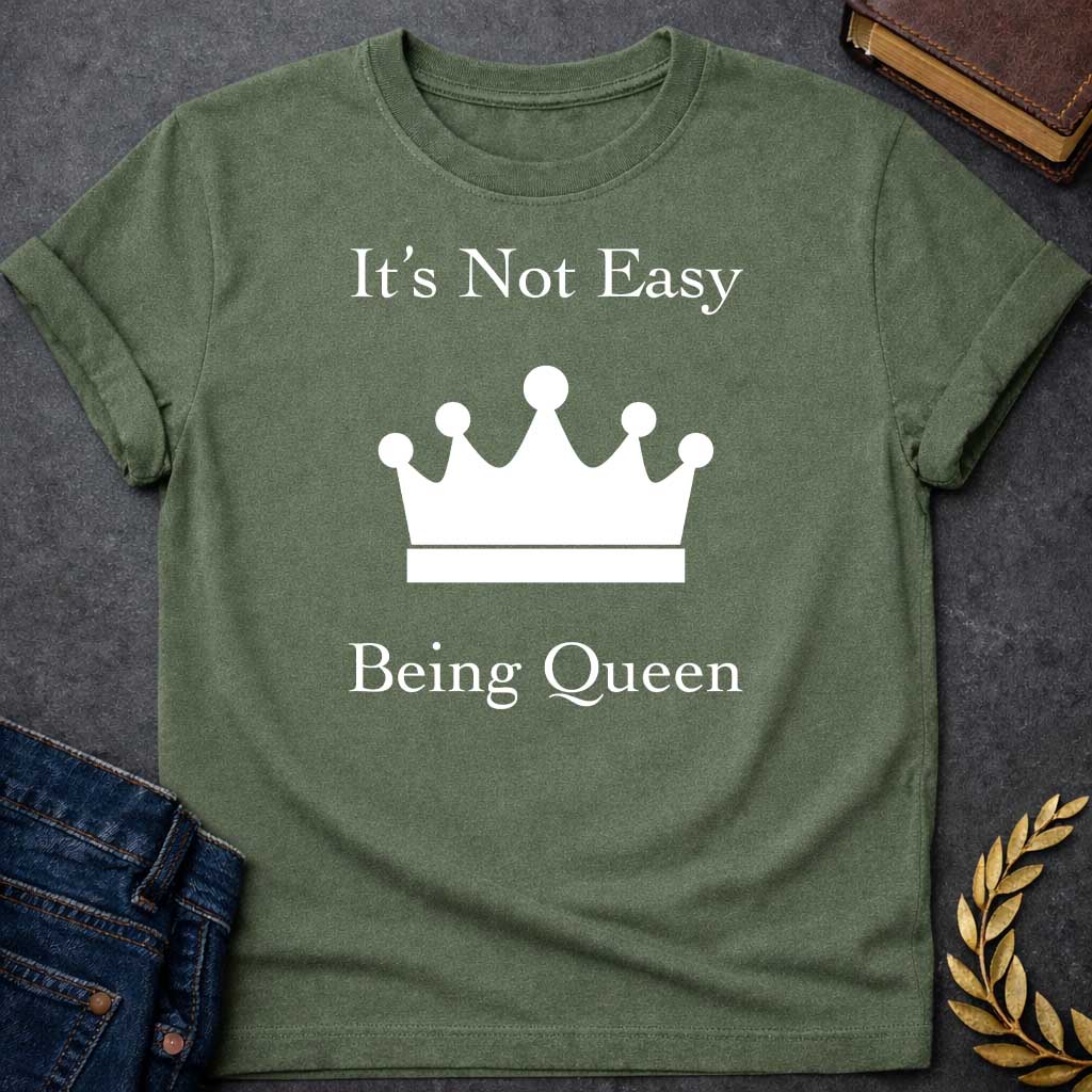 Military Green T-shirt graphic in white on black featuring elegant serif text reading "It's Not Easy Being Queen" split above and below a simple white crown silhouette icon, in a humorous nod to Hera, the Greek Queen of the Gods