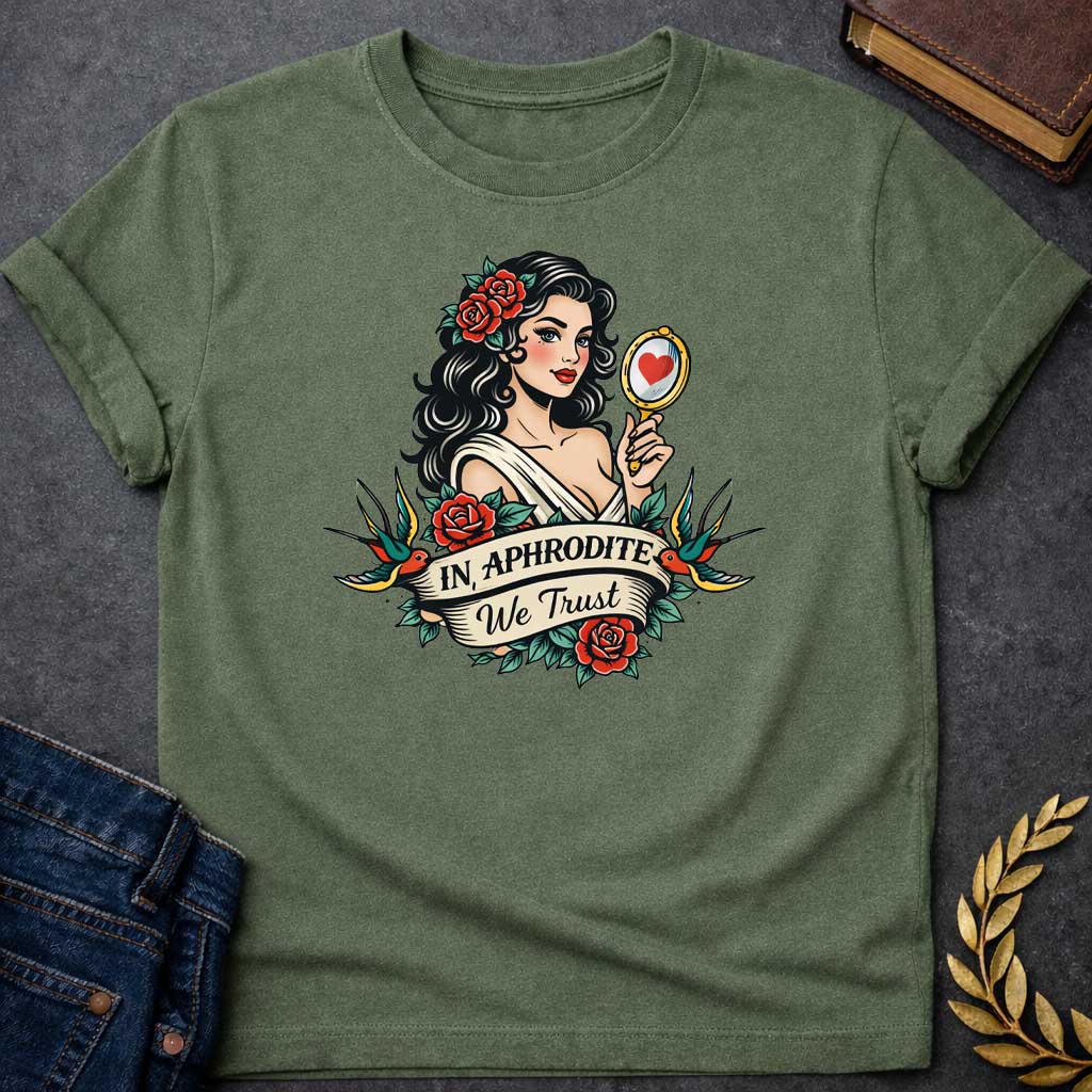 Military Green T-shirt graphic in a classic American traditional tattoo style featuring a pin-up illustration of Aphrodite, the Greek Goddess of Love, with dark wavy hair adorned with red roses, wearing a white draped gown, holding a gold hand mirror reflecting a red heart. She is flanked by two swooping swallows and surrounded by red roses and green leaves, with a scroll banner below reading "In Aphrodite We Trust" in bold and script lettering