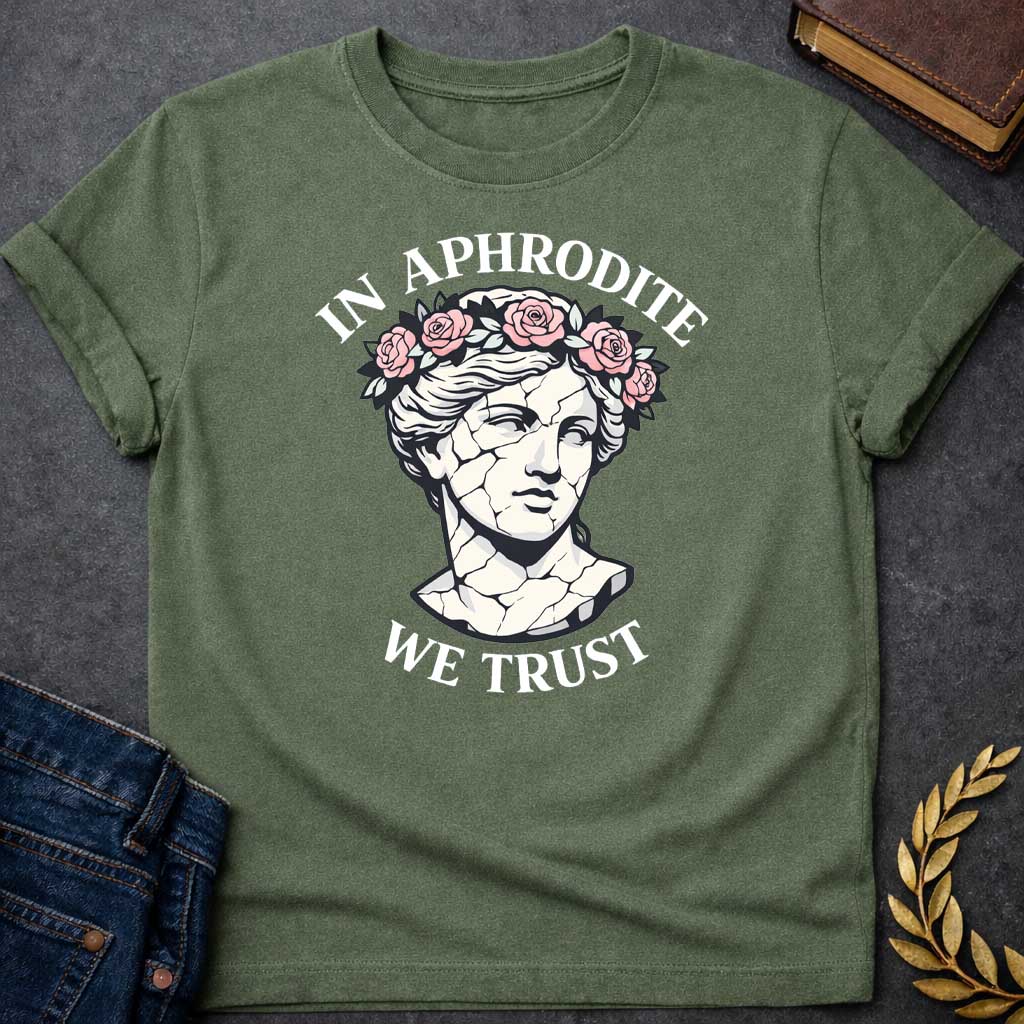 Military Green T-shirt graphic featuring a cracked marble bust illustration of Aphrodite, the Greek Goddess of Love, adorned with a floral crown of pink roses and green leaves, with bold arched text reading "In Aphrodite" curved above and "We Trust" curved below, in a modern aesthetic vaporwave-inspired style with white text and soft pink and white tones