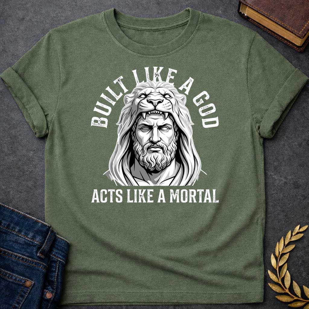 Military Green T-Shirt with a high-contrast, black-and-white graphic. The illustration depicts a stylized, statuesque man wearing the head and mane of a lion as a hood, reminiscent of Hercules. The text "BUILT LIKE A GOD" is arched over the top, and "ACTS LIKE A MORTAL" is printed in bold, straight text across the botto