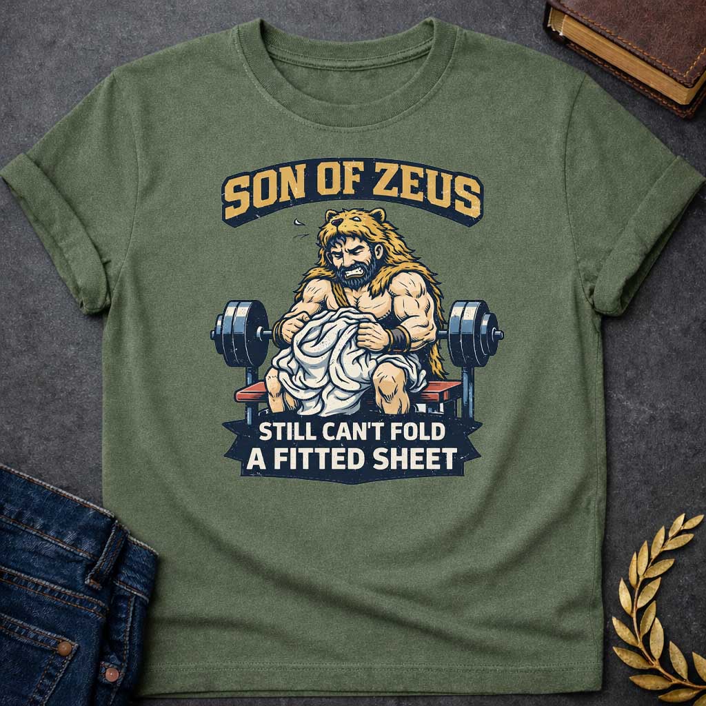 Military Green T-shirt featuring a humorous illustration of a muscular, bearded man wearing a lion pelt—resembling Hercules—sitting on a weight bench. He is struggling, with a frustrated expression, to fold a crumpled white fitted sheet. Behind him is a barbell. The text at the top reads "SON OF ZEUS" in a distressed, arched font, and the text at the bottom reads "STILL CAN'T FOLD A FITTED SHEET" in a banner