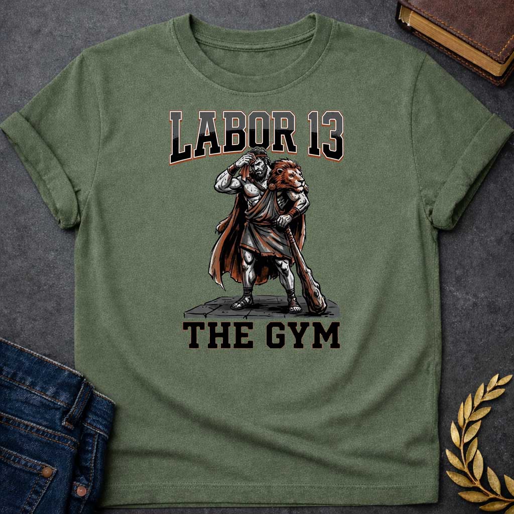 Military Green T-shirt featuring a sweat-drenched, muscular figure resembling Heracles, wearing a lion-skin cloak and holding a large wooden club. He stands on a stone platform while wiping his brow with a towel. The text "LABOR 13" is printed in large, bold, block letters across the top, and "THE GYM" is in a matching style at the bottom