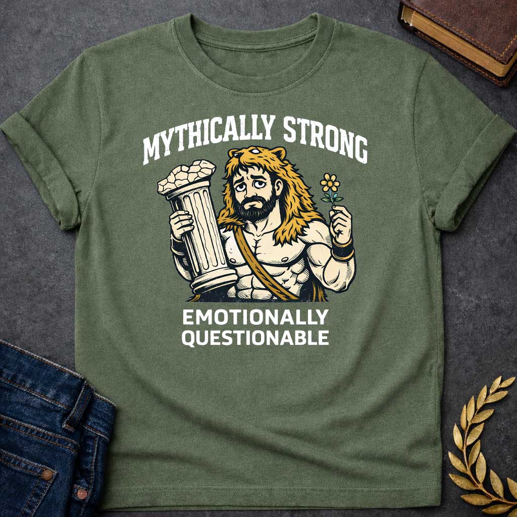 Military Green T-shirt featuring a humorous illustration of a muscular, bearded man wearing a lion-skin hood—reminiscent of Hercules—with a sad, weary expression. He holds a broken classical column in one hand and a small flower in the other. Arched over the top is the white text "MYTHICALLY STRONG," and below the illustration is the text "EMOTIONALLY QUESTIONABLE,"