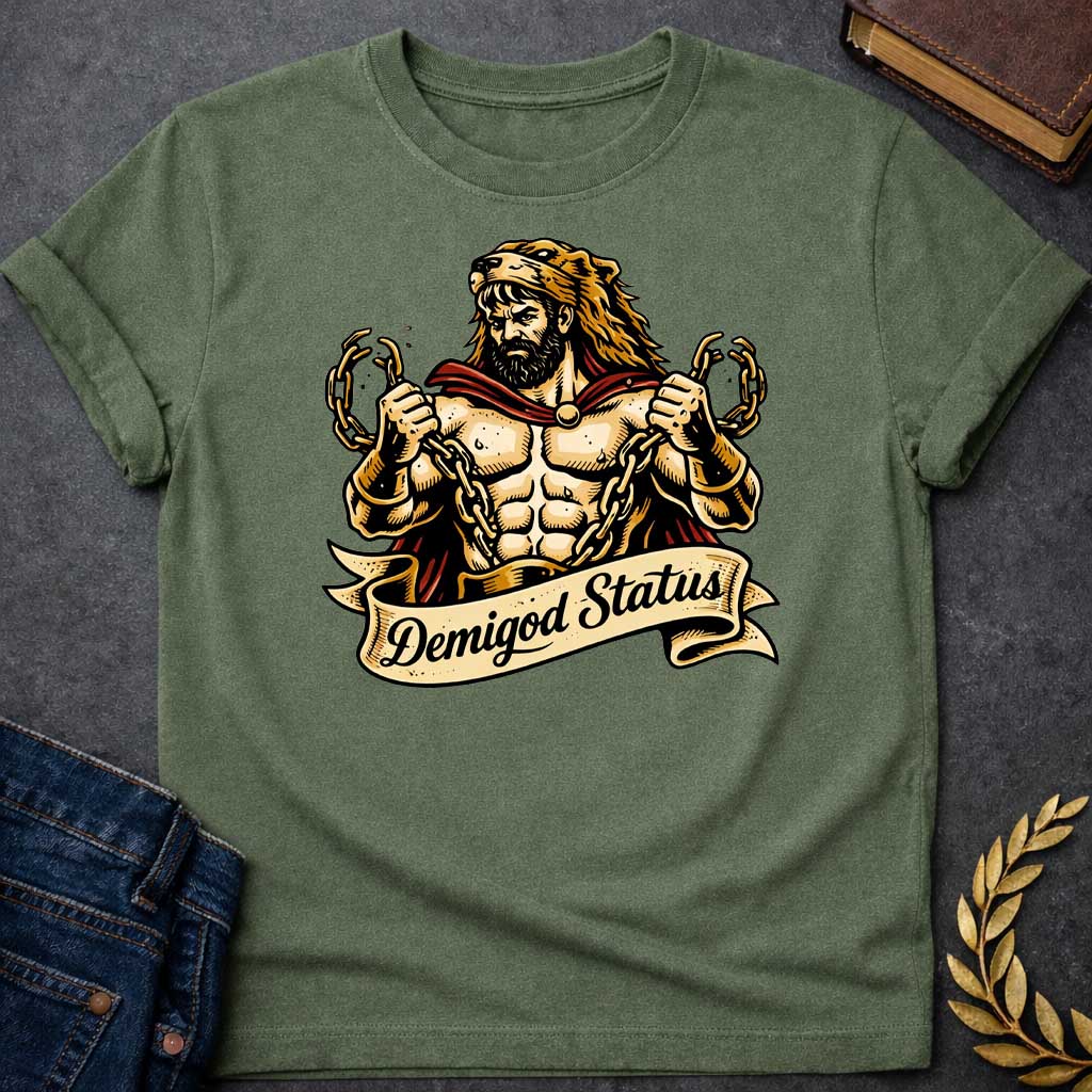 Military Green T-shirt featuring a muscular, bearded figure resembling Heracles, wearing a lion-skin hood and a red cape, as he heroically snaps thick golden chains with his hands. Below the illustration is a flowing, decorative banner with the text "Demigod Status" written in a stylish script font,