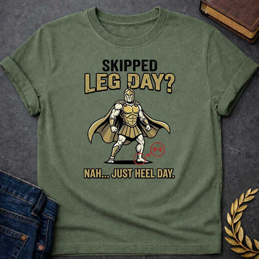 Military Green T-Shirt with a humorous cartoon combining Greek mythology with gym culture. A muscular Greek warrior in heroic stance wears gold Corinthian helmet, armor with defined abs, pleated skirt, and cape. The warrior has an exaggerated upper body but the left heel is circled in red with an arrow pointing to a barbell icon. Text reads 'SKIPPED LEG DAY?' above and 'NAH... JUST HEEL DAY' below. Comedic mashup referencing Achilles' vulnerable heel and the modern fitness joke about skipping leg day.