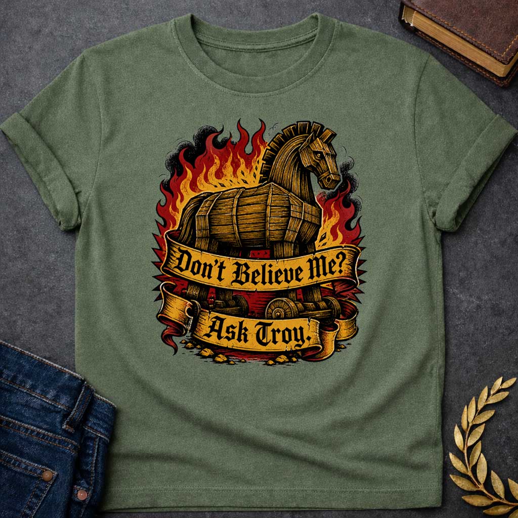 Military Green T-shirt with a bold illustration of the Trojan Horse engulfed in flames with red, orange, and yellow fire and black smoke. The wooden horse emerges from flames with detailed wood-grain texture. Two ornate gold banners with blackletter text read 'Don't Believe Me?' and 'Ask Troy.' Dark red background with rubble scattered below. References Cassandra's ignored warning about the Trojan Horse that led to Troy's destruction. Dramatic tattoo-inspired style with vintage engraving detail.