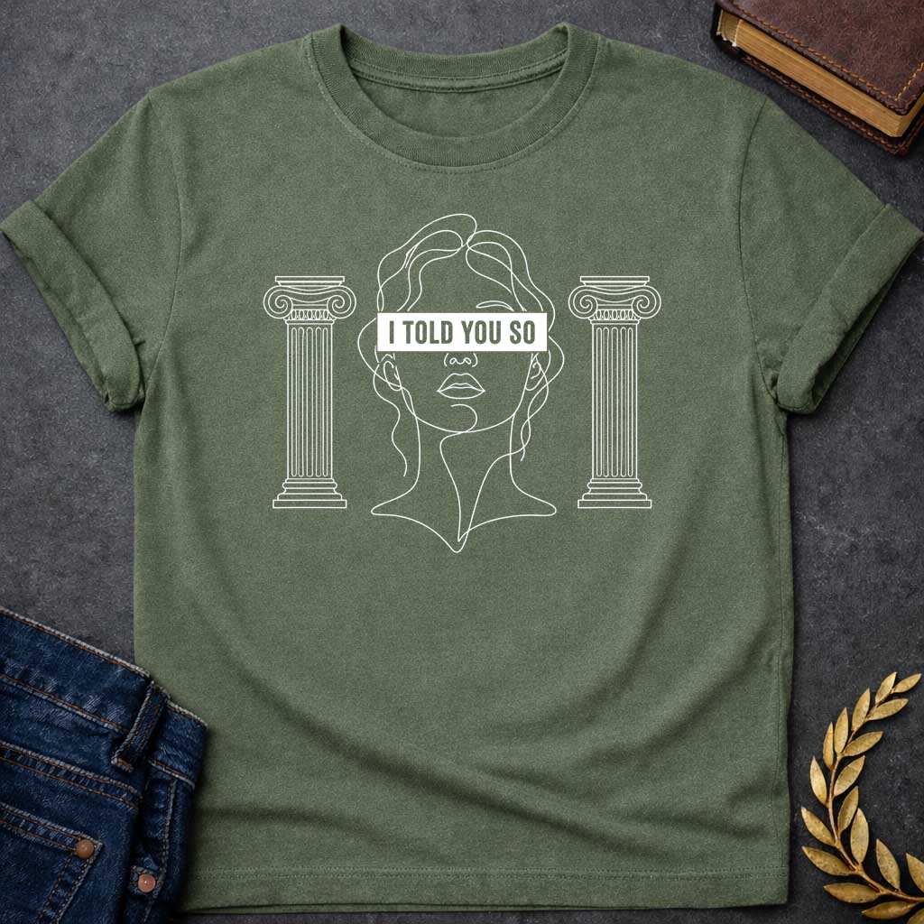Military Green T-shirt with a minimalist line art design featuring a continuous white ink sketch of Cassandra or a Greek goddess. Her eyes are covered by a bold, solid black rectangular bar with the text "I TOLD YOU SO" in white sans-serif capital letters. The portrait is flanked on both sides by simple, symmetrical line-drawn Ionic Greek columns.