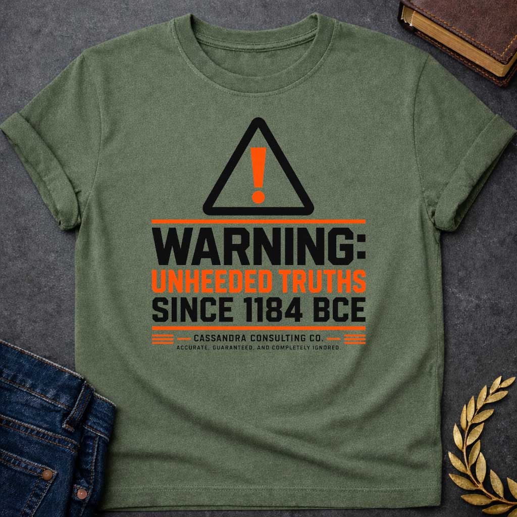 Military Green T-shirt with a corporate-style satirical design mimicking an industrial hazard sign. At the top is a triangular warning icon with an orange exclamation point. Bold block text reads "WARNING: UNHEEDED TRUTHS SINCE 1184 BCE" in alternating black and orange. Below, it features a faux company name: "CASSANDRA CONSULTING CO." followed by the tagline, "ACCURATE, GUARANTEED, AND COMPLETELY IGNORED."
