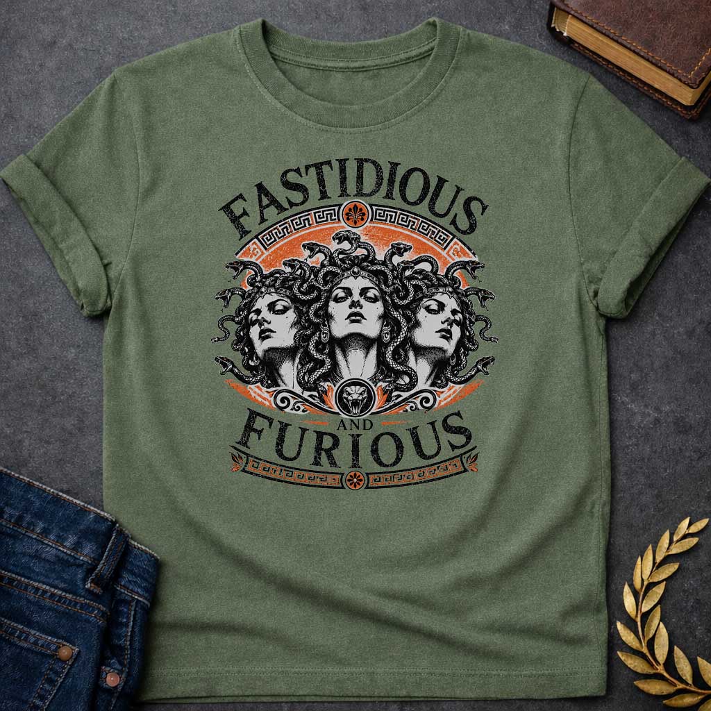 Military Green T-shirt with a Humorous design combining 'Fast and Furious' with Greek mythology. Text reads 'FASTIDIOUS AND FURIOUS' in distressed lettering surrounding three faces of Medusa in black and white engraving style. Detailed serpents with individual scales form her hair. Greek key pattern borders in orange frame the composition with decorative sun motifs and a small snake head icon at center. Blends classical mythology with contemporary pop culture humor and clever wordplay.