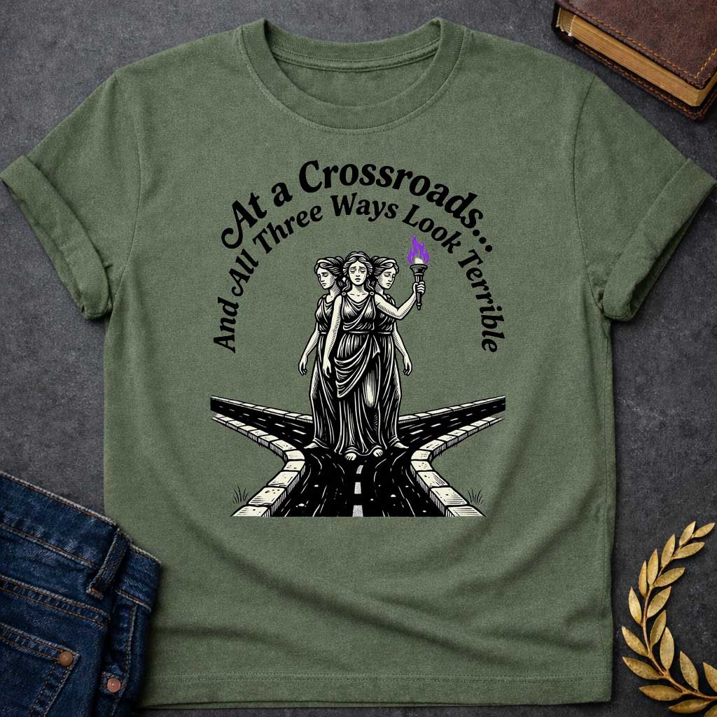 Military Green T-shirt with an Illustration of the Hecate at a crossroads, rendered in black and white engraving style. The figures wear flowing robes and stand together, with the center holding a flaming torch with purple flames. Text reads 'At a Crossroads: And All Three Ways Look Terrible,' humorously referencing difficult choices and Hecate’s role as goddess of the crossroads. Multiple roads branch beneath them in perspective, receding into darkness symbolizing uncertainty.