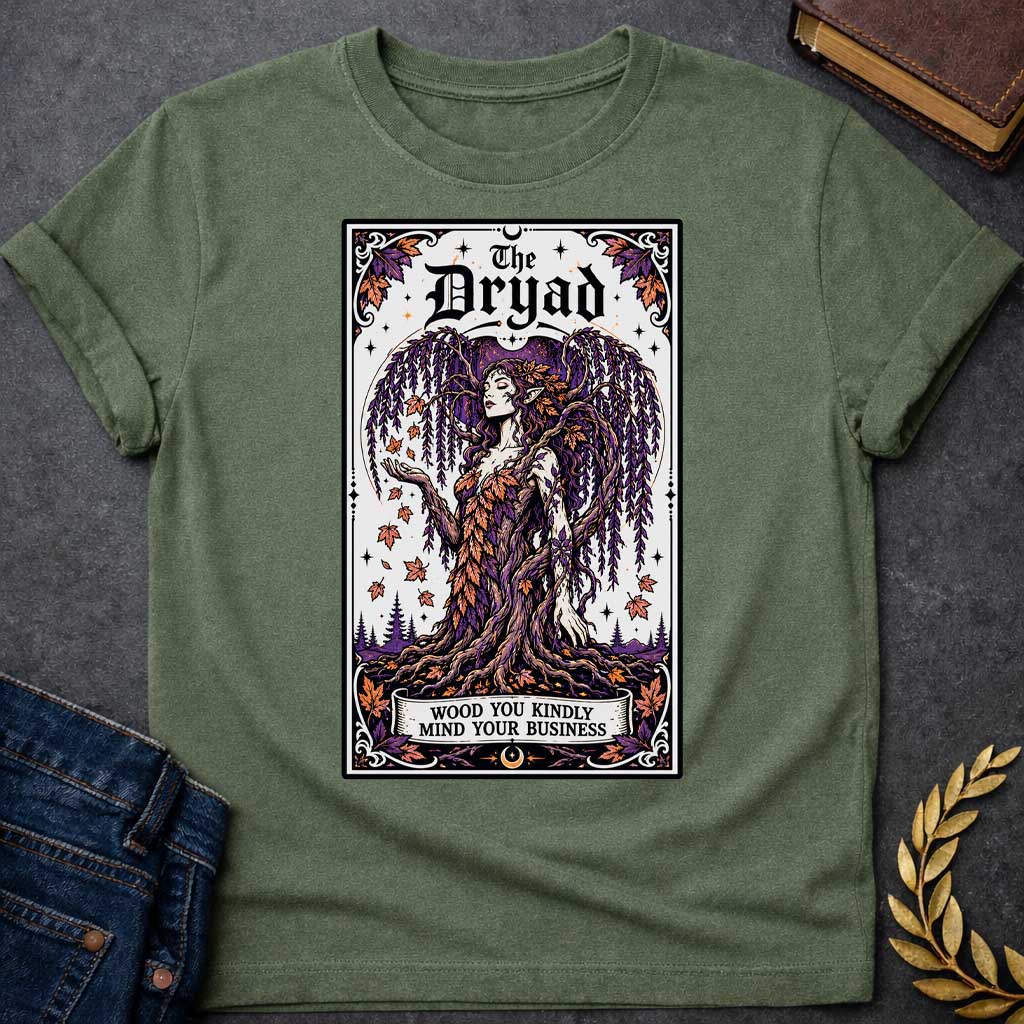 Military Green T-shirt with a mystical, tarot-card style design framed by an ornate border. It features a majestic weeping willow dryad whose gown is woven from purple and orange autumn leaves. At the top, bold gothic text reads "The Dryad." A weathered banner at the base of her rooted trunk features a clever, pun-based tagline that reads: "WOOD YOU KINDLY MIND YOUR BUSINESS" in a clean, black serif font