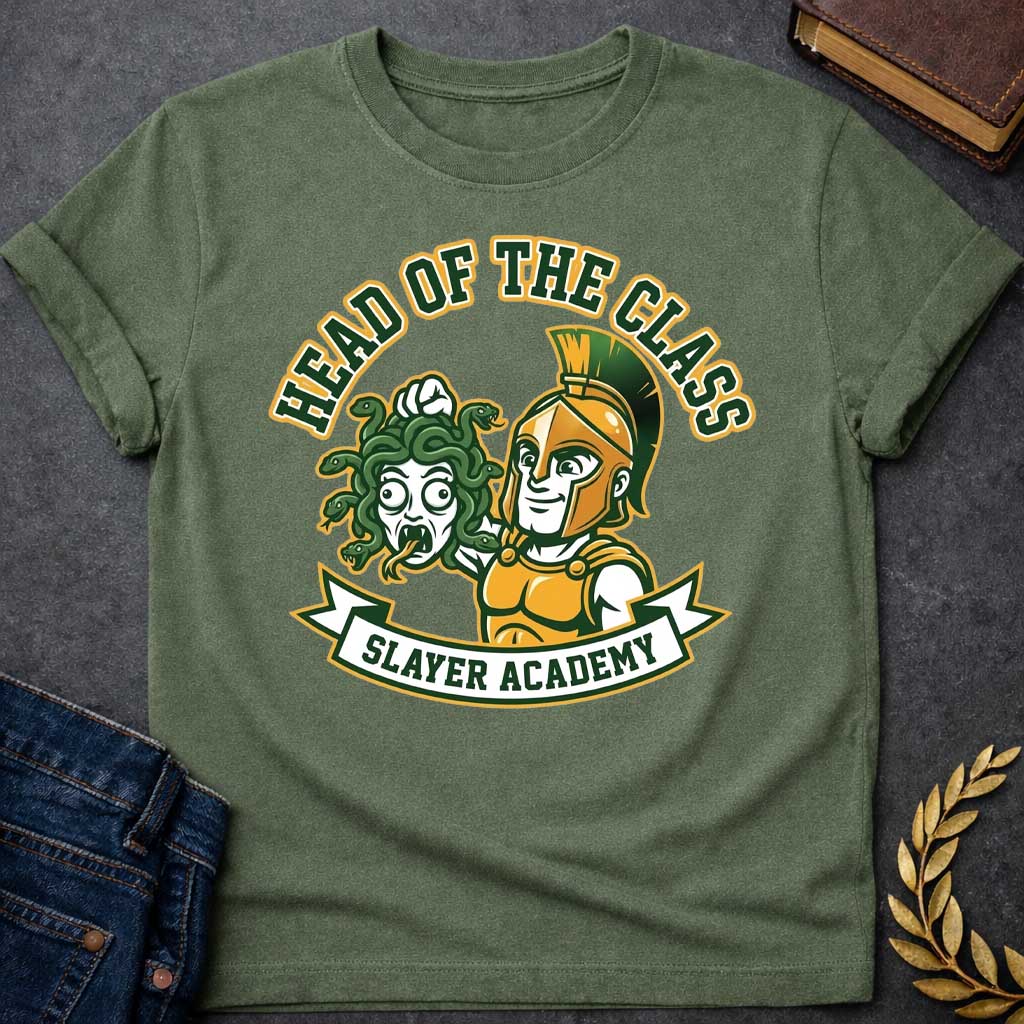 Military Green t-shirt in collegiate mascot style: a smirking gold-helmeted Greek warrior holds up Medusa's shocked, snarling severed head by her green snake hair. Bold green and gold text arcs above the design, with "Slayer Academy" on a white ribbon banner below. Playful varsity-logo aesthetic on a white background.