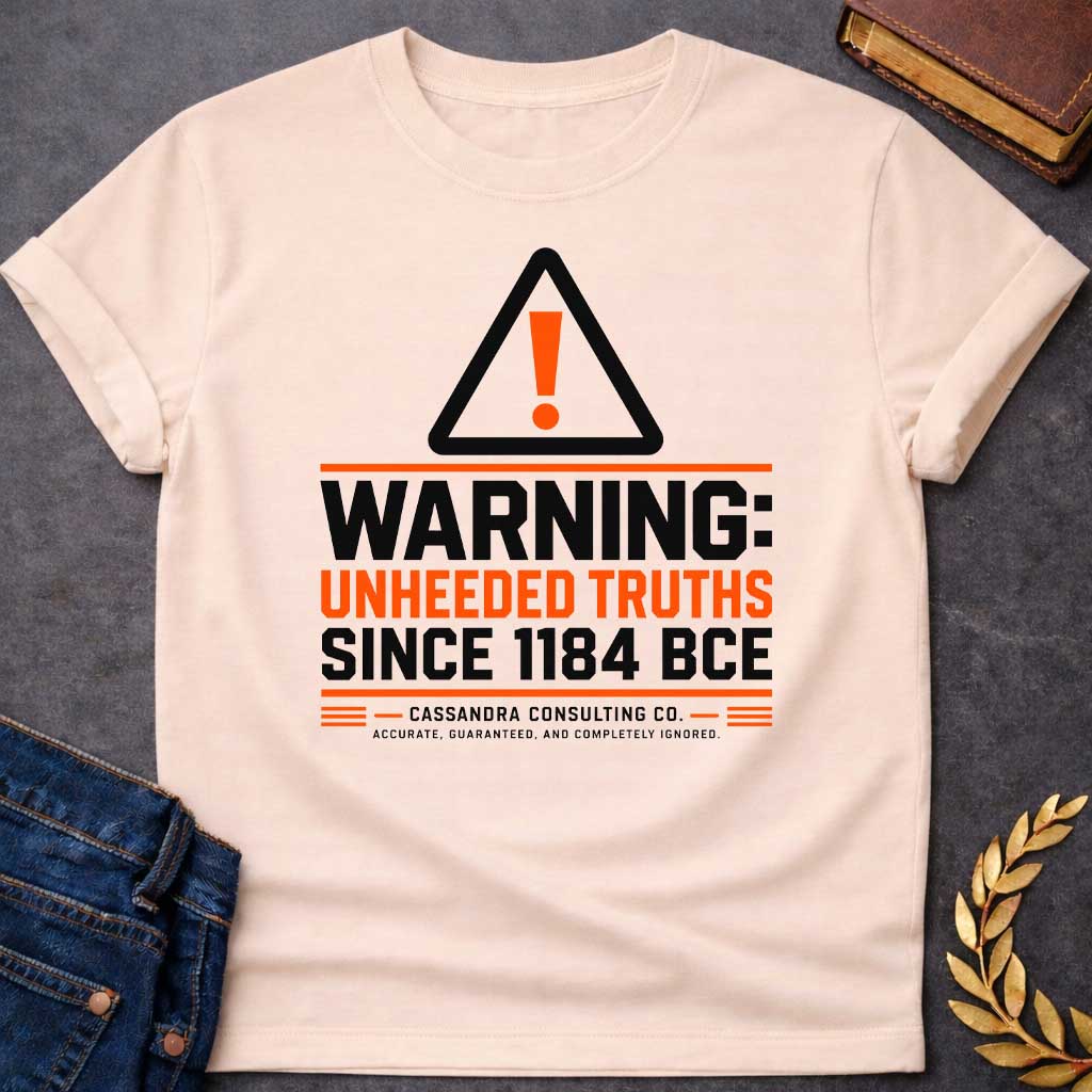 Natural T-shirt with a corporate-style satirical design mimicking an industrial hazard sign. At the top is a triangular warning icon with an orange exclamation point. Bold block text reads "WARNING: UNHEEDED TRUTHS SINCE 1184 BCE" in alternating black and orange. Below, it features a faux company name: "CASSANDRA CONSULTING CO." followed by the tagline, "ACCURATE, GUARANTEED, AND COMPLETELY IGNORED."