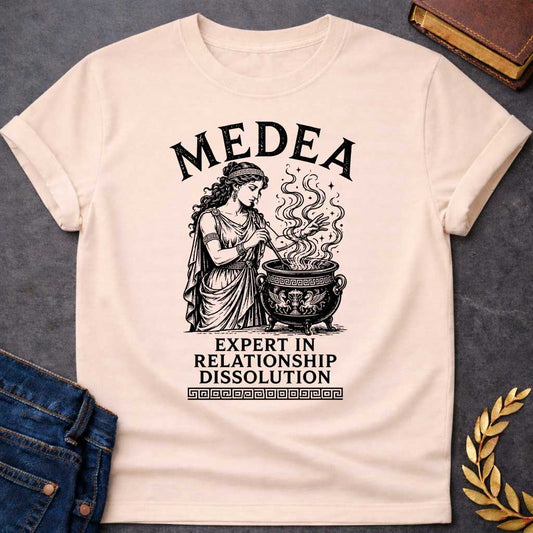 Natural T-shirt with a Classical engraving of Medea, the legendary Greek sorceress, in flowing draped robes with Greek key pattern headband. She stirs a decorative cauldron with magical swirling smoke and stars rising dramatically from within. Black and white line art style with intricate detailing. Text reads 'Medea' above and 'Expert in Relationship Dissolution' below with Greek key border. Dark humor celebrates Medea's infamous reputation for magical vengeance and relationship destruction.