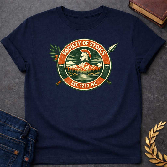 Navy T-shirt graphic featuring a circular vintage badge design with the text "Society of Stoics" arched above and "Est. 1217 BC" below. The center illustration shows a Spartan helmet floating above a mountain landscape with pine trees and a reflective lake, rendered in dark green, orange, and cream tones. Two crossed spears extend behind the badge, with an olive branch decorating the upper left.