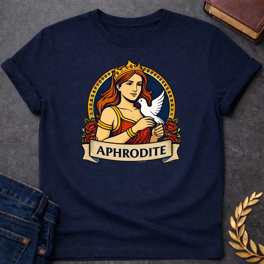 Navy T-shirt graphic featuring a vibrant illustrated portrait of Aphrodite, the Greek Goddess of Love, with flowing red hair and a gold crown, wearing a red draped gown with gold bracelets, cradling a white dove in her hands. She is framed within a gold circular border flanked by red roses, with a scroll banner below reading "Aphrodite," in a bold tattoo-inspired style with navy, gold, and red tones
