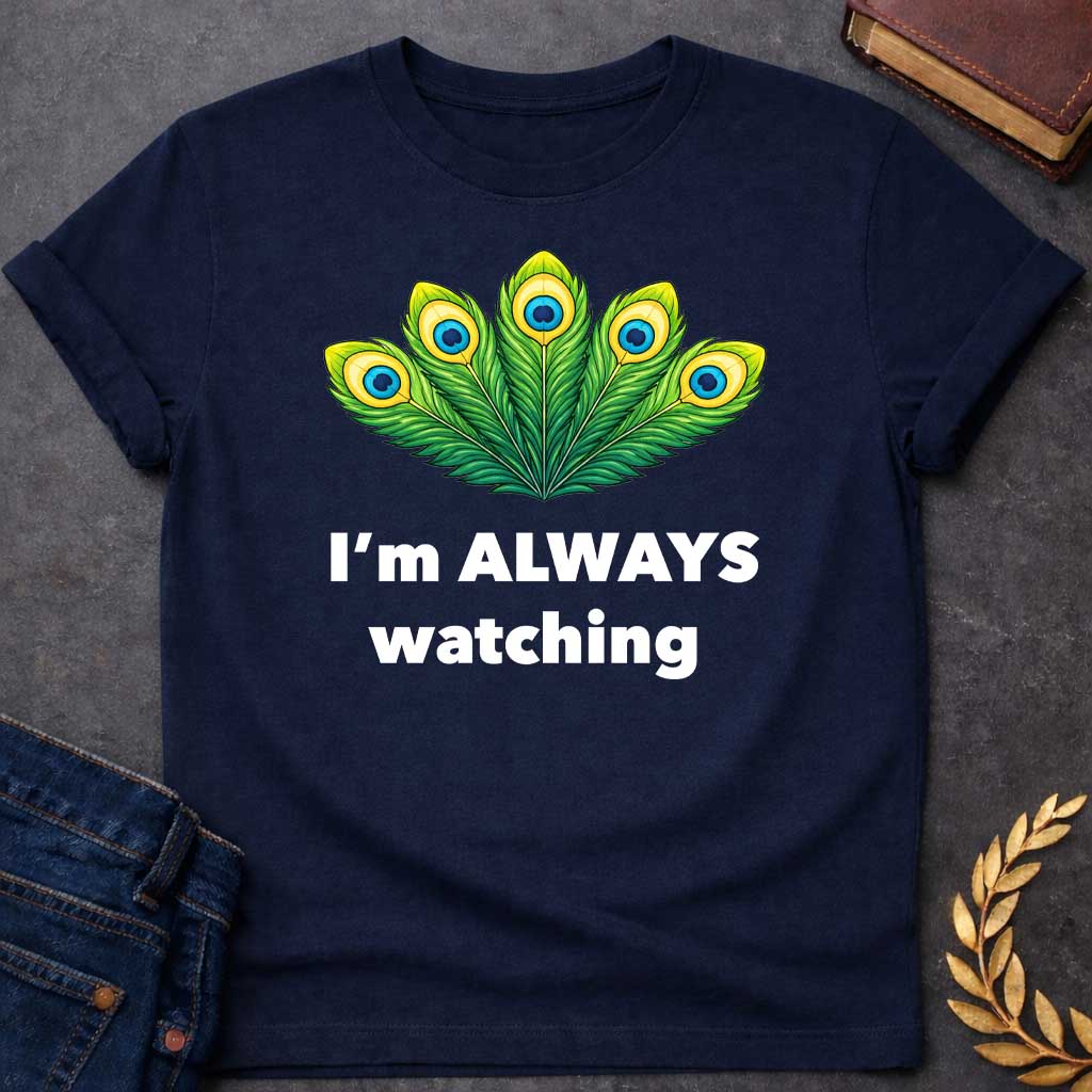 Navy T-shirt graphic featuring a vibrant illustrated fan of five peacock feathers in rich green, yellow, and blue tones, each with a distinctive eye-spot, above text reading "I'm Always Watching" — a humorous reference to Hera, the Greek Queen of the Gods, whose sacred symbol is the peacock
