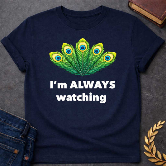 Navy T-shirt graphic featuring a vibrant illustrated fan of five peacock feathers in rich green, yellow, and blue tones, each with a distinctive eye-spot, above text reading "I'm Always Watching" — a humorous reference to Hera, the Greek Queen of the Gods, whose sacred symbol is the peacock