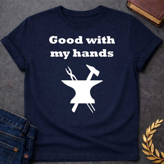 Navy T-shirt graphic in white featuring bold serif text reading "Good with my hands" above a flat black silhouette illustration of a blacksmith's anvil with a hammer and tongs crossed on top — a humorous nod to Hephaestus, the Greek God of the Forge