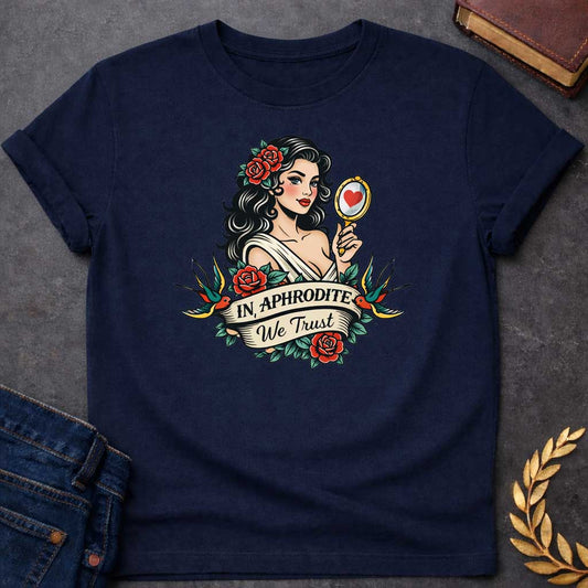 Navy T-shirt graphic in a classic American traditional tattoo style featuring a pin-up illustration of Aphrodite, the Greek Goddess of Love, with dark wavy hair adorned with red roses, wearing a white draped gown, holding a gold hand mirror reflecting a red heart. She is flanked by two swooping swallows and surrounded by red roses and green leaves, with a scroll banner below reading "In Aphrodite We Trust" in bold and script lettering