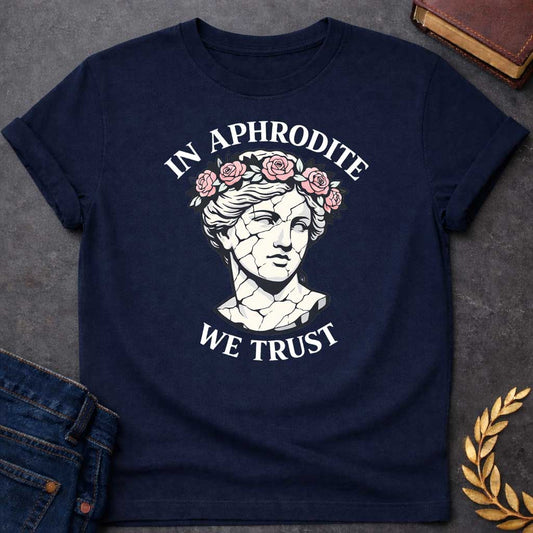 Navy T-shirt graphic featuring a cracked marble bust illustration of Aphrodite, the Greek Goddess of Love, adorned with a floral crown of pink roses and green leaves, with bold arched text reading "In Aphrodite" curved above and "We Trust" curved below, in a modern aesthetic vaporwave-inspired style with white text and soft pink and white tones