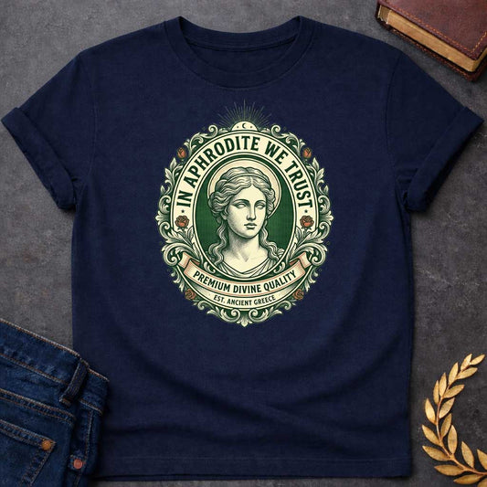 Navy T-shirt graphic featuring a vintage apothecary label-style oval badge design in forest green, cream, and terracotta. A detailed engraving-style portrait of Aphrodite is centered within the badge, surrounded by ornate scrollwork and flanked by red roses, with a crescent moon and sunburst radiating from the top. Text arched around the inner circle reads "In Aphrodite We Trust," with a scroll banner below reading "Premium Divine Quality" and smaller text underneath reading "Est. Ancient Greece,"