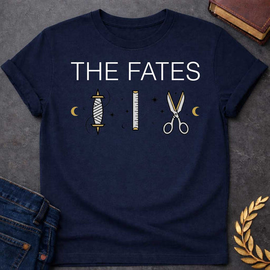Navy T-shirt graphic featuring large bold white text reading "The Fates" above three minimalist icons representing the tools of the Moirai, the Greek Fates — a spindle with thread, a measuring ruler, and a pair of scissors — each surrounded by subtle crescent moons and celestial star details, rendered in white and gold tones