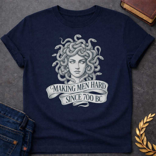 Navy T-shirt graphic in grey and white featuring a highly detailed fine-line tattoo-style front-facing portrait of Medusa with numerous coiling snakes for hair, rendered in intricate stippling and engraving technique, with a flowing ribbon banner below reading "Making Men Hard Since 700 BC" — a witty double entendre referencing Medusa's mythological power to turn men to stone