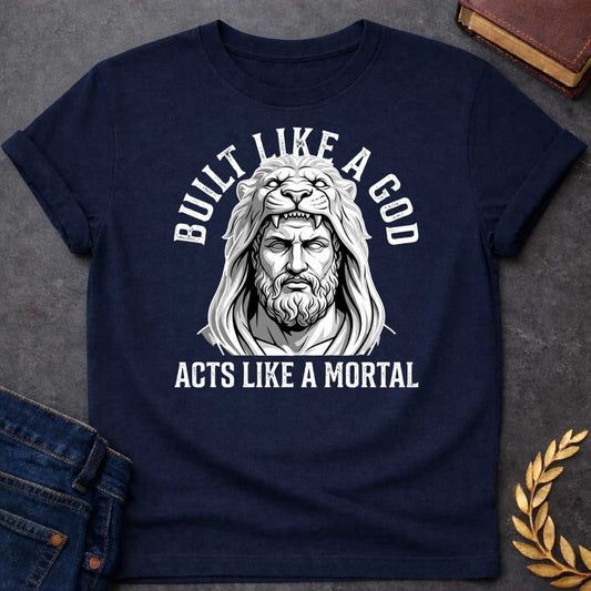 Navy T-Shirt with a high-contrast, black-and-white graphic. The illustration depicts a stylized, statuesque man wearing the head and mane of a lion as a hood, reminiscent of Hercules. The text "BUILT LIKE A GOD" is arched over the top, and "ACTS LIKE A MORTAL" is printed in bold, straight text across the botto