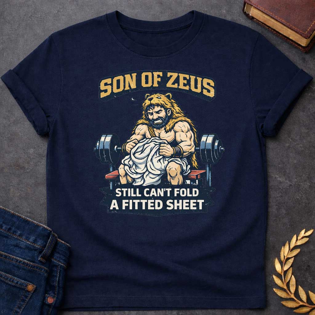 Navy T-shirt featuring a humorous illustration of a muscular, bearded man wearing a lion pelt—resembling Hercules—sitting on a weight bench. He is struggling, with a frustrated expression, to fold a crumpled white fitted sheet. Behind him is a barbell. The text at the top reads "SON OF ZEUS" in a distressed, arched font, and the text at the bottom reads "STILL CAN'T FOLD A FITTED SHEET" in a banner