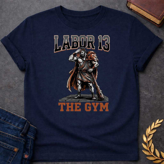 Navy T-shirt featuring a sweat-drenched, muscular figure resembling Heracles, wearing a lion-skin cloak and holding a large wooden club. He stands on a stone platform while wiping his brow with a towel. The text "LABOR 13" is printed in large, bold, block letters across the top, and "THE GYM" is in a matching style at the bottom