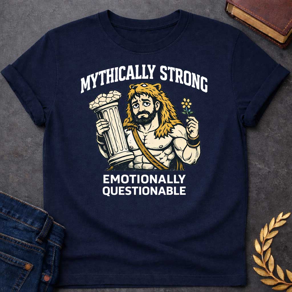Navy T-shirt featuring a humorous illustration of a muscular, bearded man wearing a lion-skin hood—reminiscent of Hercules—with a sad, weary expression. He holds a broken classical column in one hand and a small flower in the other. Arched over the top is the white text "MYTHICALLY STRONG," and below the illustration is the text "EMOTIONALLY QUESTIONABLE,"