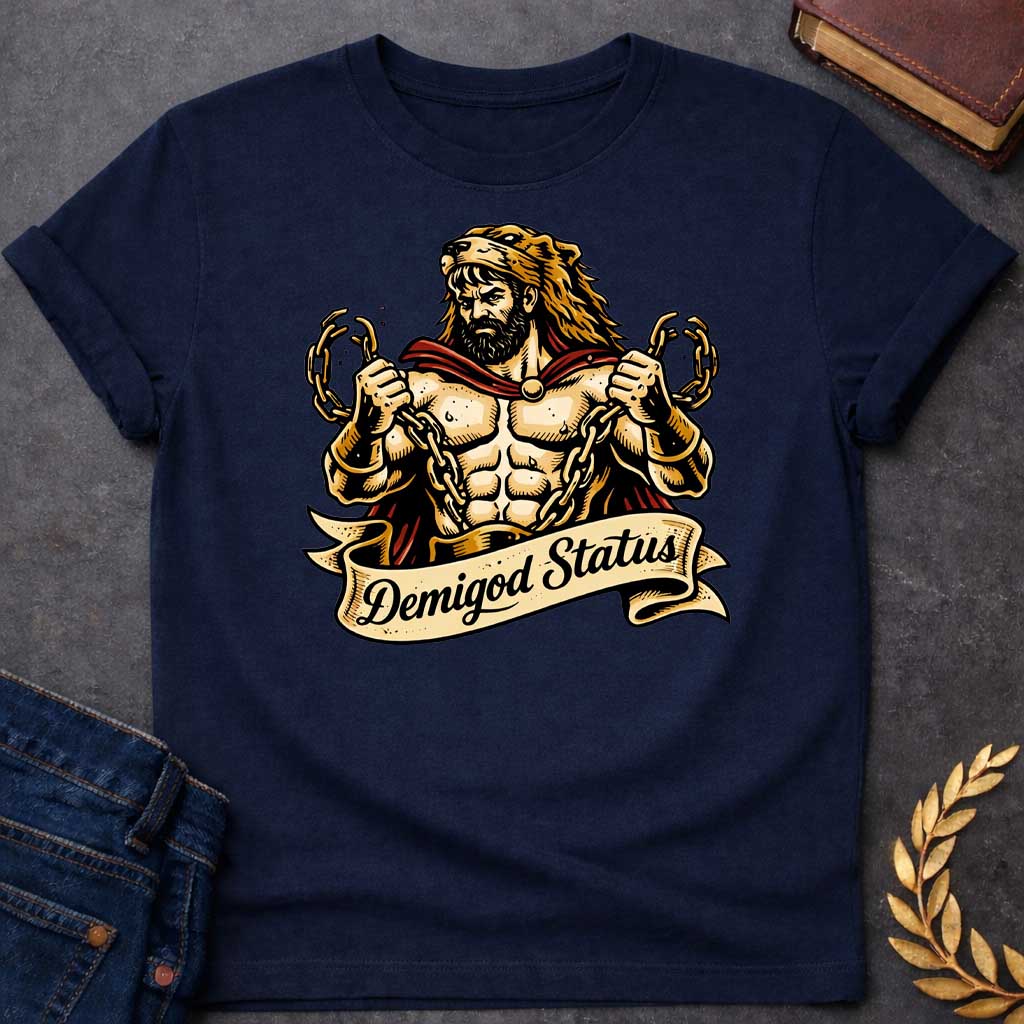 Navy T-shirt featuring a muscular, bearded figure resembling Heracles, wearing a lion-skin hood and a red cape, as he heroically snaps thick golden chains with his hands. Below the illustration is a flowing, decorative banner with the text "Demigod Status" written in a stylish script font,