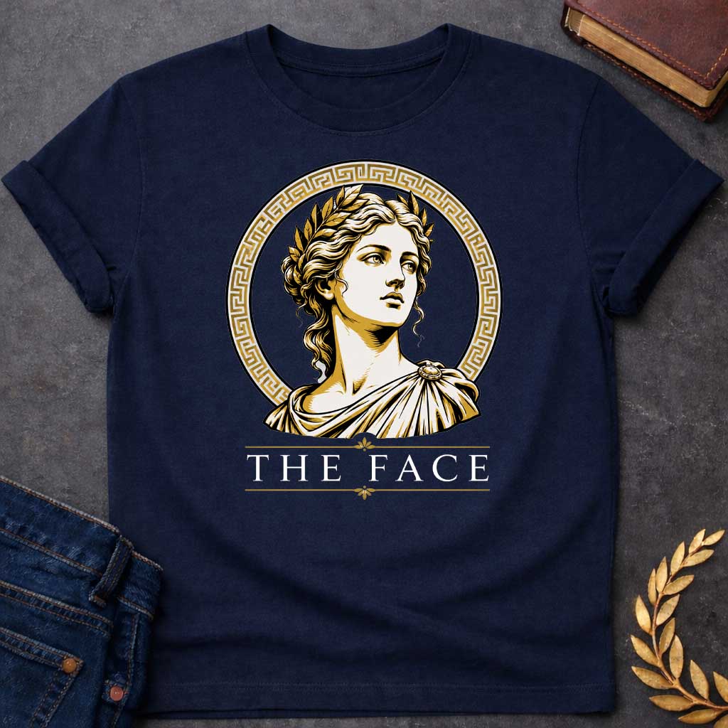 Navy T-shirt with a classical Greek graphic featuring a gold and cream vector portrait of Helen of Troy in profile. She wears a golden laurel wreath and a traditional draped chiton, looking upward with a serene expression. The portrait is framed by a circular border with a gold Greek key (meander) pattern. Below the emblem, the text "THE FACE" is written in a clean, elegant white serif font between horizontal gold accent lines.