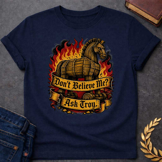 Navy T-shirt with a bold illustration of the Trojan Horse engulfed in flames with red, orange, and yellow fire and black smoke. The wooden horse emerges from flames with detailed wood-grain texture. Two ornate gold banners with blackletter text read 'Don't Believe Me?' and 'Ask Troy.' Dark red background with rubble scattered below. References Cassandra's ignored warning about the Trojan Horse that led to Troy's destruction. Dramatic tattoo-inspired style with vintage engraving detail.