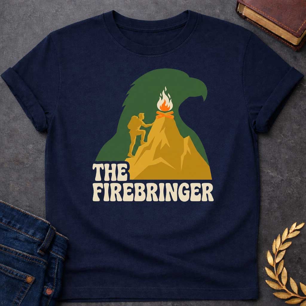 Navy T-shirt with a retro, outdoor-style t-shirt graphic referencing Prometheus. It features a silhouette of a hiker with a backpack climbing a golden mountain peak toward a campfire at the summit. The entire mountain scene is set against a large background silhouette of an eagle's head in forest green. Below the image, the text "THE FIREBRINGER" is written in a bold, stylized 1970s-style cream font.