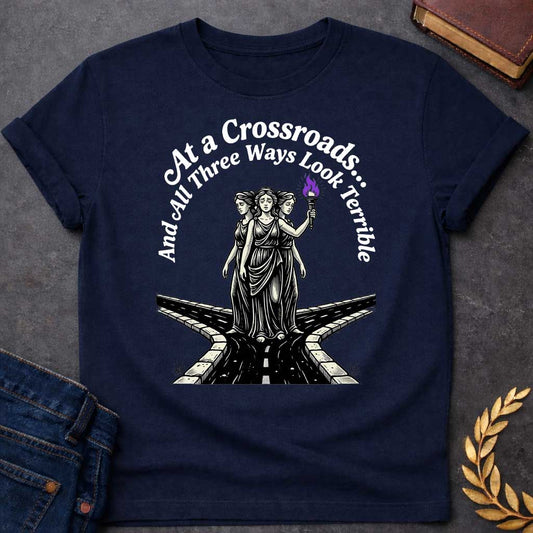Navy T-shirt with an Illustration of the Hecate at a crossroads, rendered in black and white engraving style. The figures wear flowing robes and stand together, with the center holding a flaming torch with purple flames. Text reads 'At a Crossroads: And All Three Ways Look Terrible,' humorously referencing difficult choices and Hecate’s role as goddess of the crossroads. Multiple roads branch beneath them in perspective, receding into darkness symbolizing uncertainty.