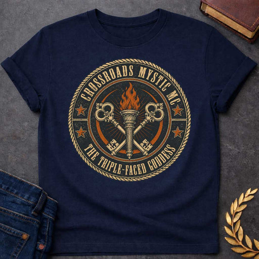 Navy T-shirt with a Motorcycle club patch-style emblem in navy, gold, and orange inspired by Hecate and crossroads mythology. A central ornate torch with orange flames is flanked by two crossed serpent handles with decorative gold scrollwork. Radiating lines suggest divine power. Text reads 'Crossroads Mystic MC' and 'The Triple-Faced Goddess' around the border. Orange stars and rope border complete the official club patch look. Distressed texture adds authentic biker aesthetic.