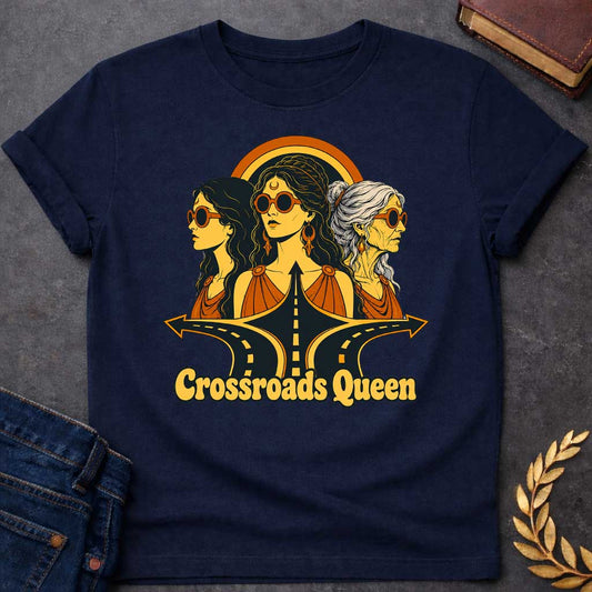 Navy T-shirt with a Glamorous retro illustration of Hecate as 'Crossroads Queen' in gold and orange with 70s aesthetic. Three female faces wear oversized round sunglasses and gold jewelry including crescent moon symbols and hoops. Center figure wears a crescent moon bindi. All wear draped orange clothing. A golden halo radiates behind them. Below, stylized black roads with yellow center lines converge at a crossroads. Text reads 'Crossroads Queen' in gold.