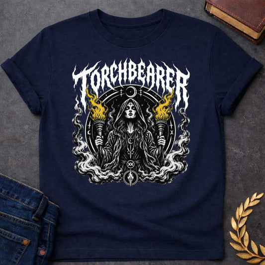Navy T-shirt with a Dark metal band aesthetic illustration titled 'Torchbearer.' Hecate holds two ornate torches with gold and black flames. She wears mystical symbols including a crescent moon necklace and a circular mandala halo with occult iconography. Swirling smoke and flame designs surround the figure in black, white, and gold. Blends heavy metal imagery with Greek mythology and mystical symbolism celebrating enlightenment, divine knowledge, and dark power.
