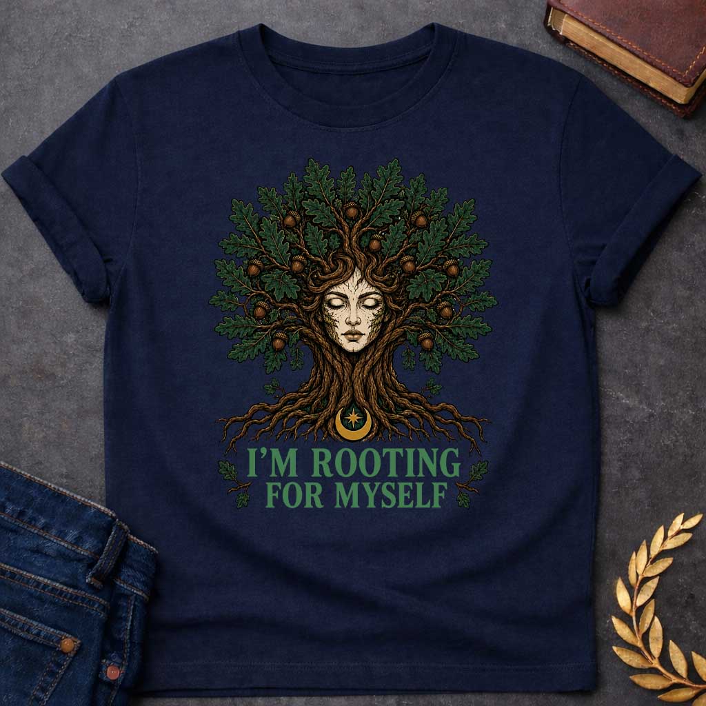 Navy T-shirt with a cottagecore-style graphic featuring a detailed illustration of an oak tree dryad or nature spirit. Her serene, closed-eyed face forms the center of the tree trunk, with gnarled roots expanding below and a massive canopy of dark green oak leaves and brown acorns spreading above. Underneath the roots, a pun-based tagline reads "I'M ROOTING FOR MYSELF" in a bold, clean green serif font.