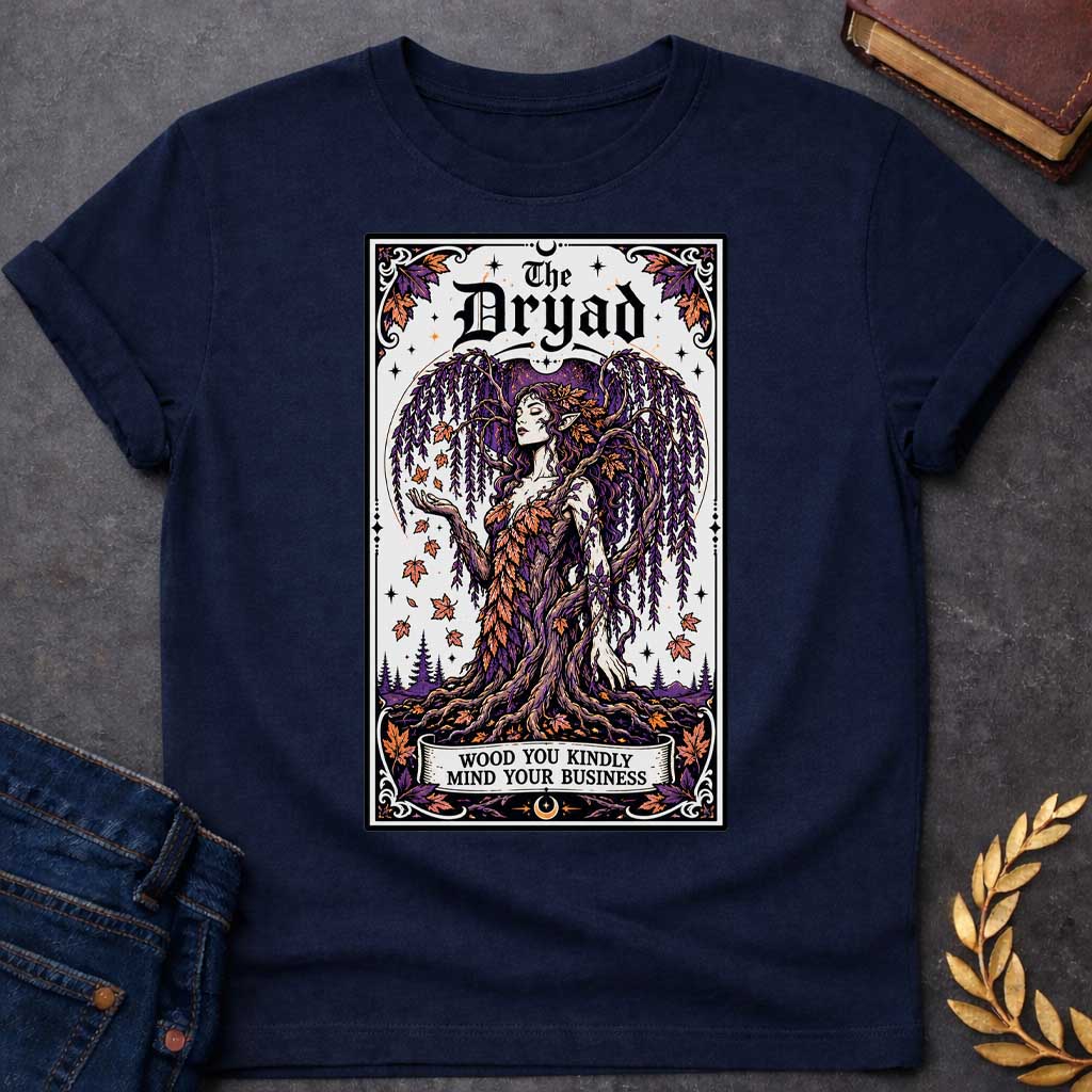 Navy T-shirt with a mystical, tarot-card style design framed by an ornate border. It features a majestic weeping willow dryad whose gown is woven from purple and orange autumn leaves. At the top, bold gothic text reads "The Dryad." A weathered banner at the base of her rooted trunk features a clever, pun-based tagline that reads: "WOOD YOU KINDLY MIND YOUR BUSINESS" in a clean, black serif font