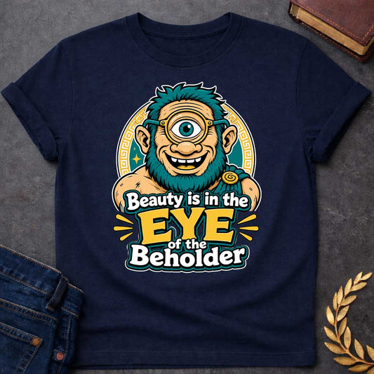 Navy T-shirt with a Humorous graphic design featuring a cheerful creature with exaggerated facial features inspired by Argus Panoptes, the many-eyed giant from Greek mythology. A large teal eye with a golden monocle dominates the character's smiling face. Teal spiky hair and beard contrast with peachy skin tones. Greek key pattern borders frame the design in gold and teal. Text reads 'Beauty is in the Eye of the Beholder' in bold white and yellow lettering. Playful visual pun celebrating perspective.