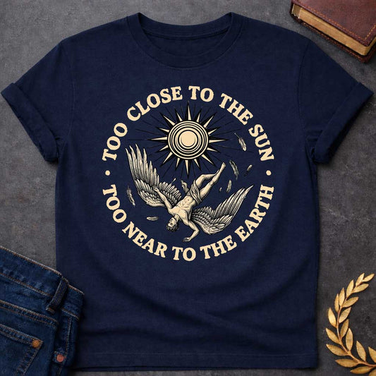 Navy t-shirt with a vintage engraved-style illustration. A winged figure of Icarus plummets downward upside down as loose feathers drift through the air beneath a large, radiating sun emblem. Encircling the detailed central artwork, bold black serif text reads "TOO CLOSE TO THE SUN" along the top arch and "TOO NEAR TO THE EARTH" along the bottom curve.