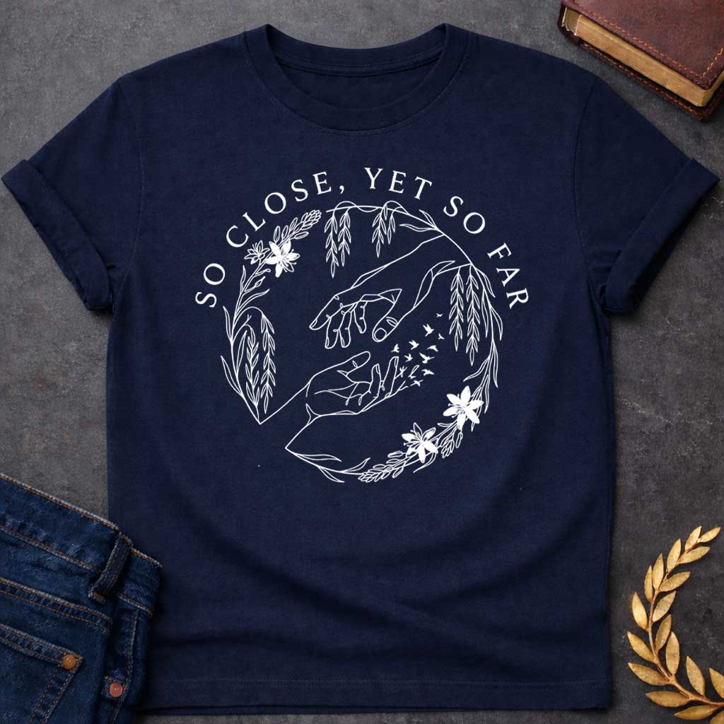 Navy t-shirt in delicate fine-line style: two reaching hands almost touch, with one hand dissolving into a flock of small birds mid-fingertip, evoking Orpheus and Eurydice. Willow branches and small flowers form a circular wreath around the scene, with elegant serif text arcing above