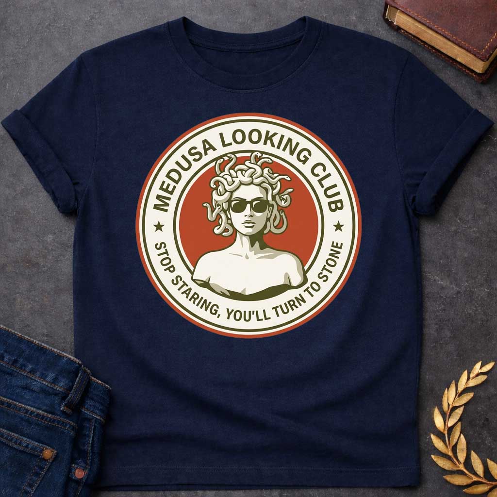 Navy t-shirt in retro badge style: a cool, sunglasses-wearing Medusa with snake hair stands confidently against a burnt-orange circle. Cream and olive-green ring text reads "Medusa Looking Club" above and "Stop Staring, You'll Turn to Stone" below, with two stars flanking the design.