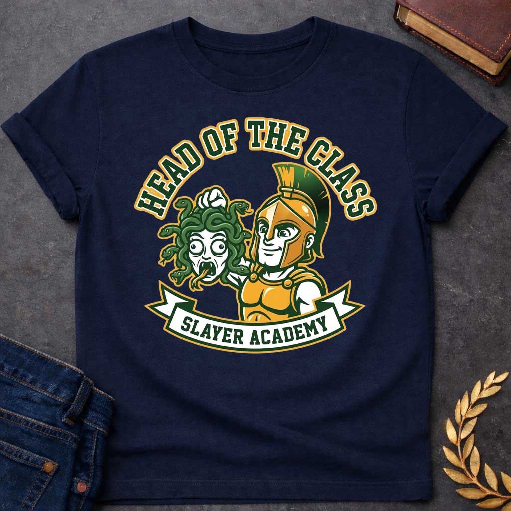 Navy t-shirt in collegiate mascot style: a smirking gold-helmeted Greek warrior holds up Medusa's shocked, snarling severed head by her green snake hair. Bold green and gold text arcs above the design, with "Slayer Academy" on a white ribbon banner below. Playful varsity-logo aesthetic on a white background.