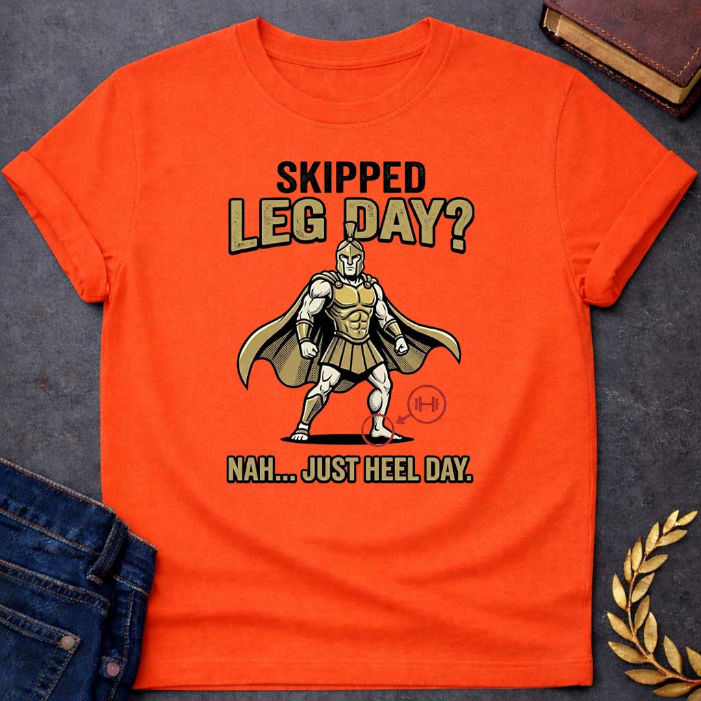 Orange T-Shirt with a humorous cartoon combining Greek mythology with gym culture. A muscular Greek warrior in heroic stance wears gold Corinthian helmet, armor with defined abs, pleated skirt, and cape. The warrior has an exaggerated upper body but the left heel is circled in red with an arrow pointing to a barbell icon. Text reads 'SKIPPED LEG DAY?' above and 'NAH... JUST HEEL DAY' below. Comedic mashup referencing Achilles' vulnerable heel and the modern fitness joke about skipping leg day.