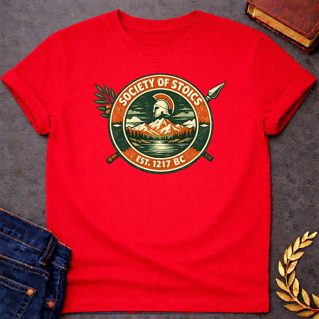 Red T-shirt graphic featuring a circular vintage badge design with the text "Society of Stoics" arched above and "Est. 1217 BC" below. The center illustration shows a Spartan helmet floating above a mountain landscape with pine trees and a reflective lake, rendered in dark green, orange, and cream tones. Two crossed spears extend behind the badge, with an olive branch decorating the upper left.