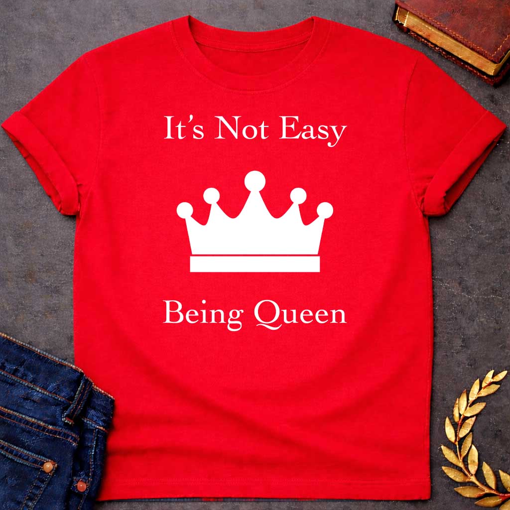 Red T-shirt graphic in white on black featuring elegant serif text reading "It's Not Easy Being Queen" split above and below a simple white crown silhouette icon, in a humorous nod to Hera, the Greek Queen of the Gods